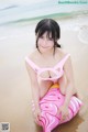 A woman in a pink bikini sitting on a pink pillow on the beach.